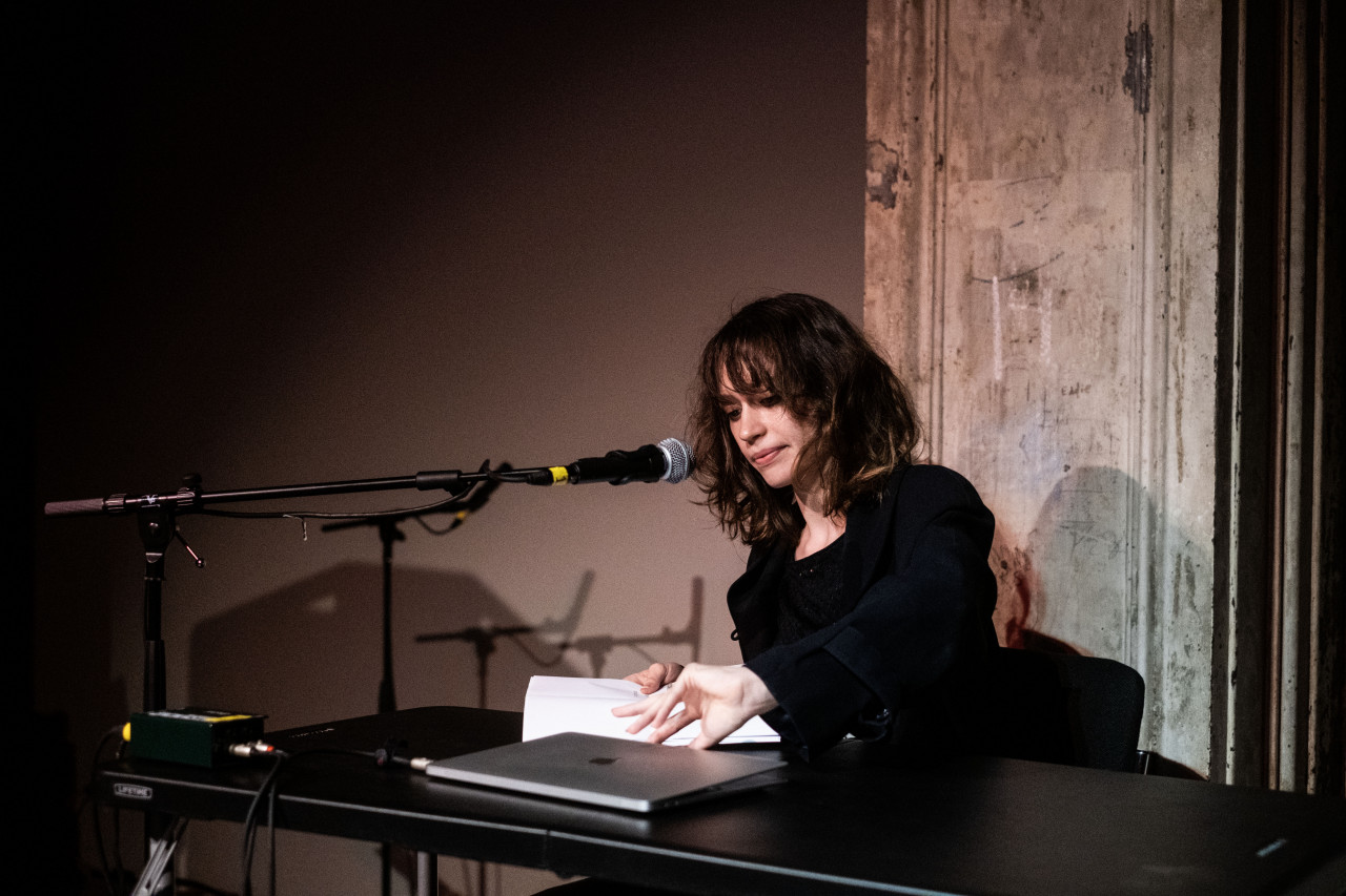 A figure sits behind a table, a microphone in front of their face, as they close their laptop and grab a stack of papers on the table.