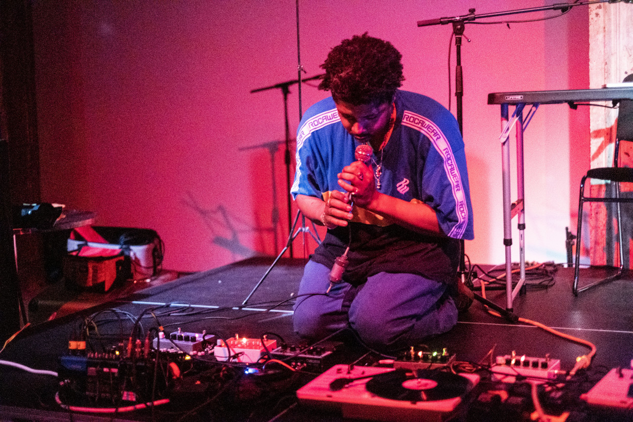 A figure in a blue shirt kneels down on stage, they look down and hold a microphone to their face. Red light is cast onto the stage.