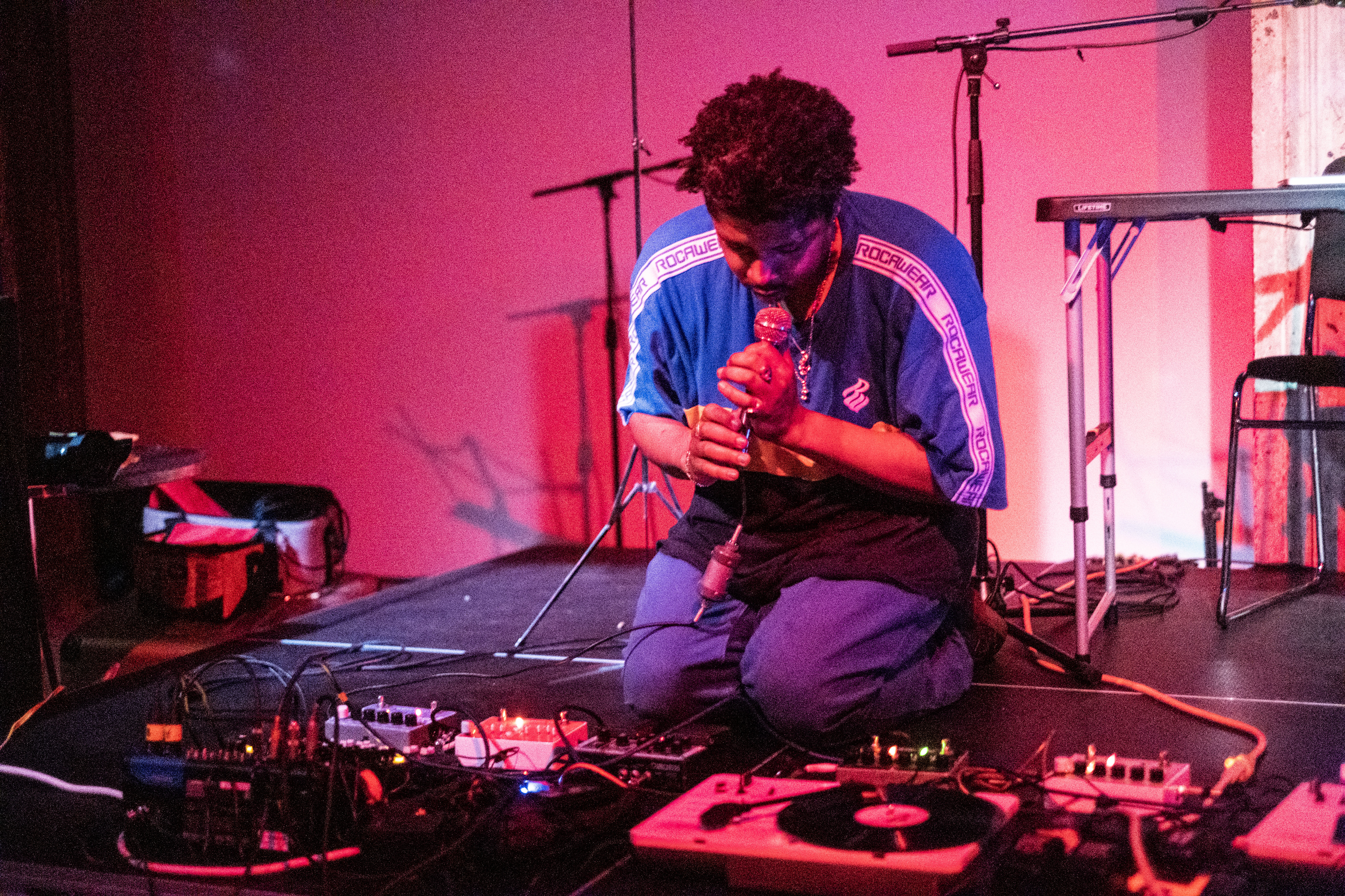 A figure in a blue shirt kneels down on stage, they look down and hold a microphone to their face. Red light is cast onto the stage.