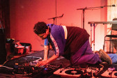 A figure kneels down on stage on all fours and sets up audio equipment layed out on the floor.