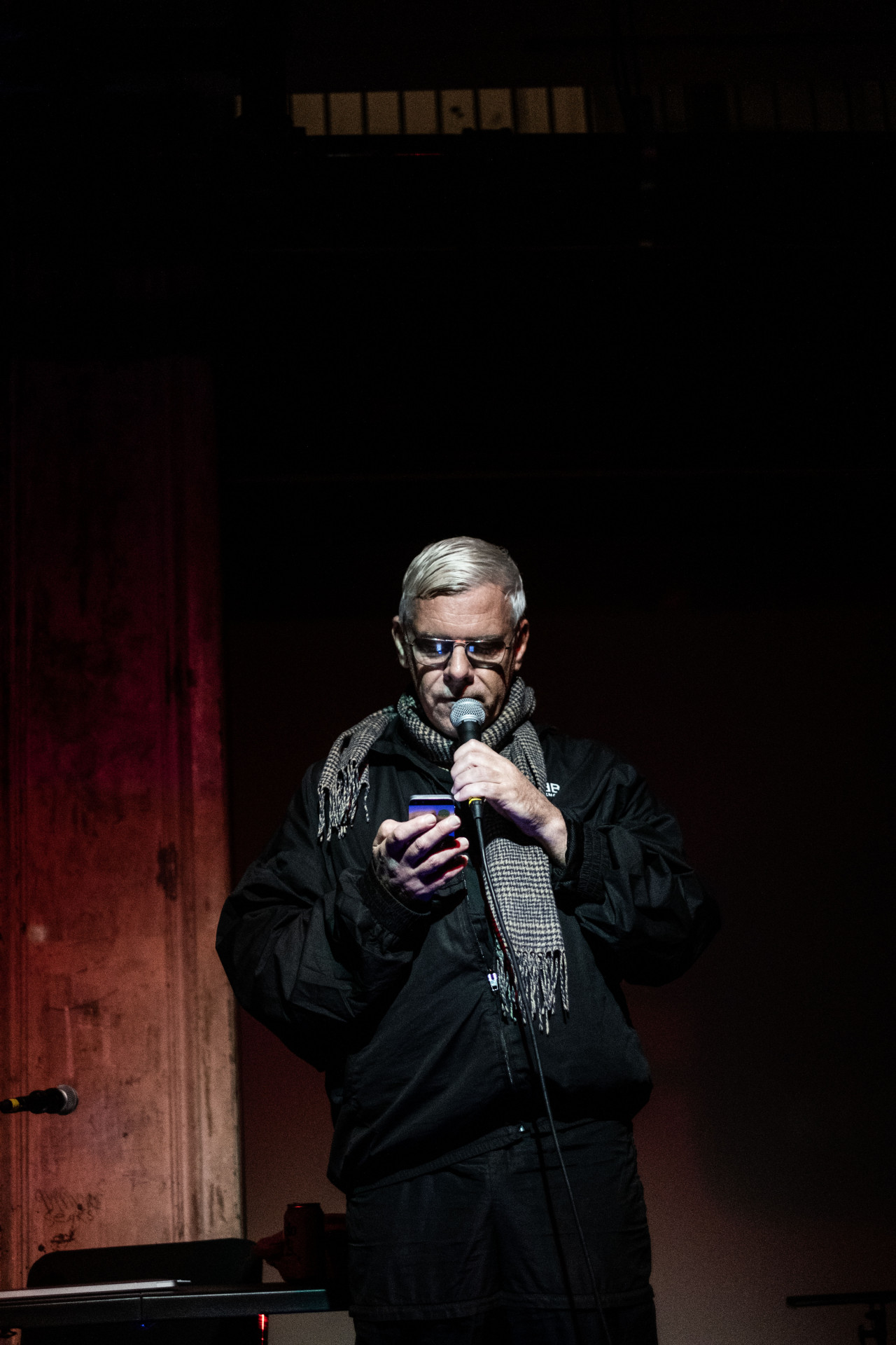 A figure reads into a microphone, glancing at their phone.