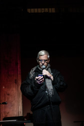 A figure reads into a microphone, glancing at their phone.