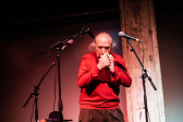 A figure in a red sweater stands on stage, with three microphones placed around them. They are blowing into a white conch shell.