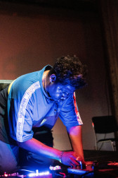 A figure in a blue shirt kneels down on stage, looking down and working with audio equipment. A blue light is cast onto them from below.