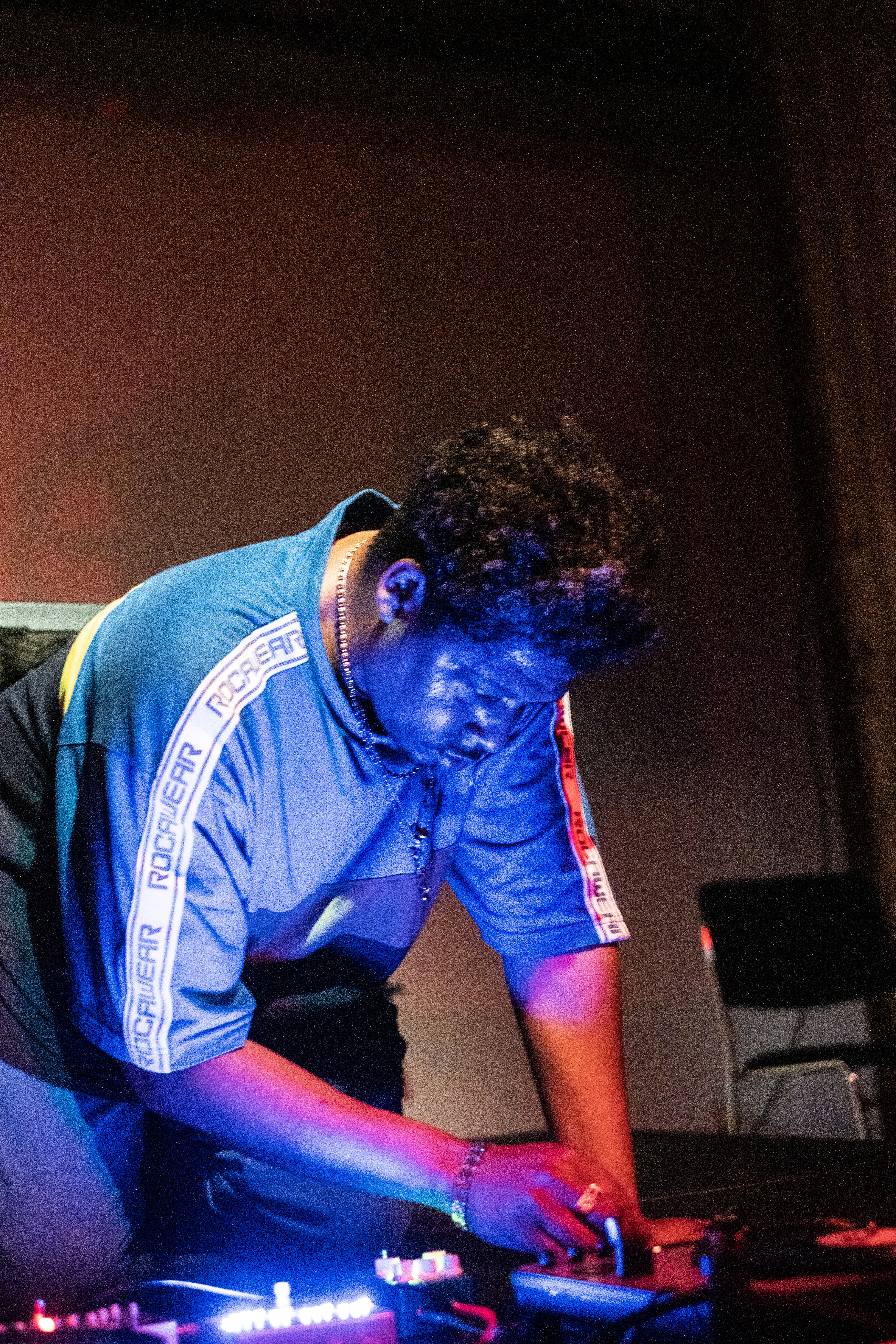 A figure in a blue shirt kneels down on stage, looking down and working with audio equipment. A blue light is cast onto them from below.