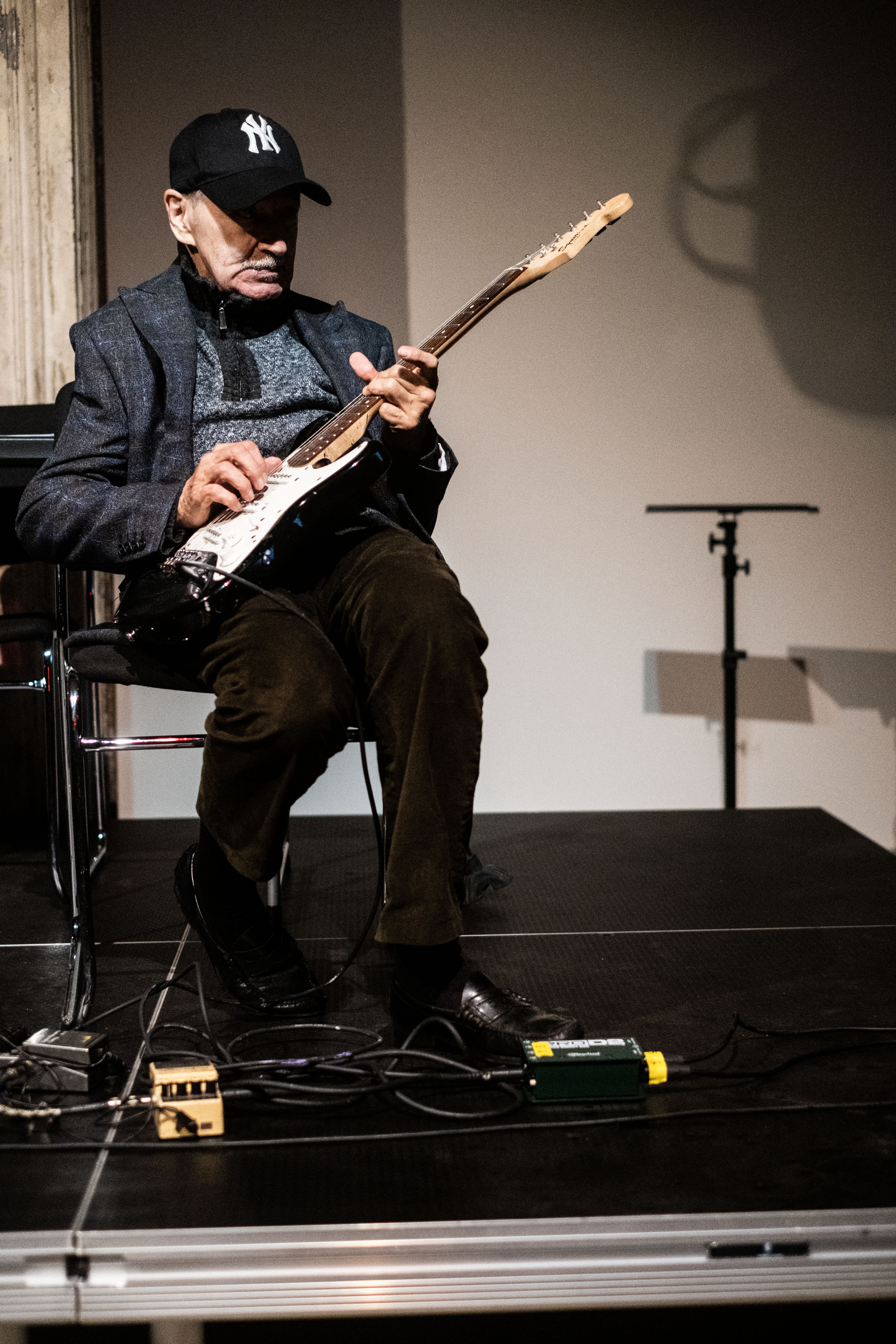 A figure wearing a baseball cap sits in a chair and plays the guitar.