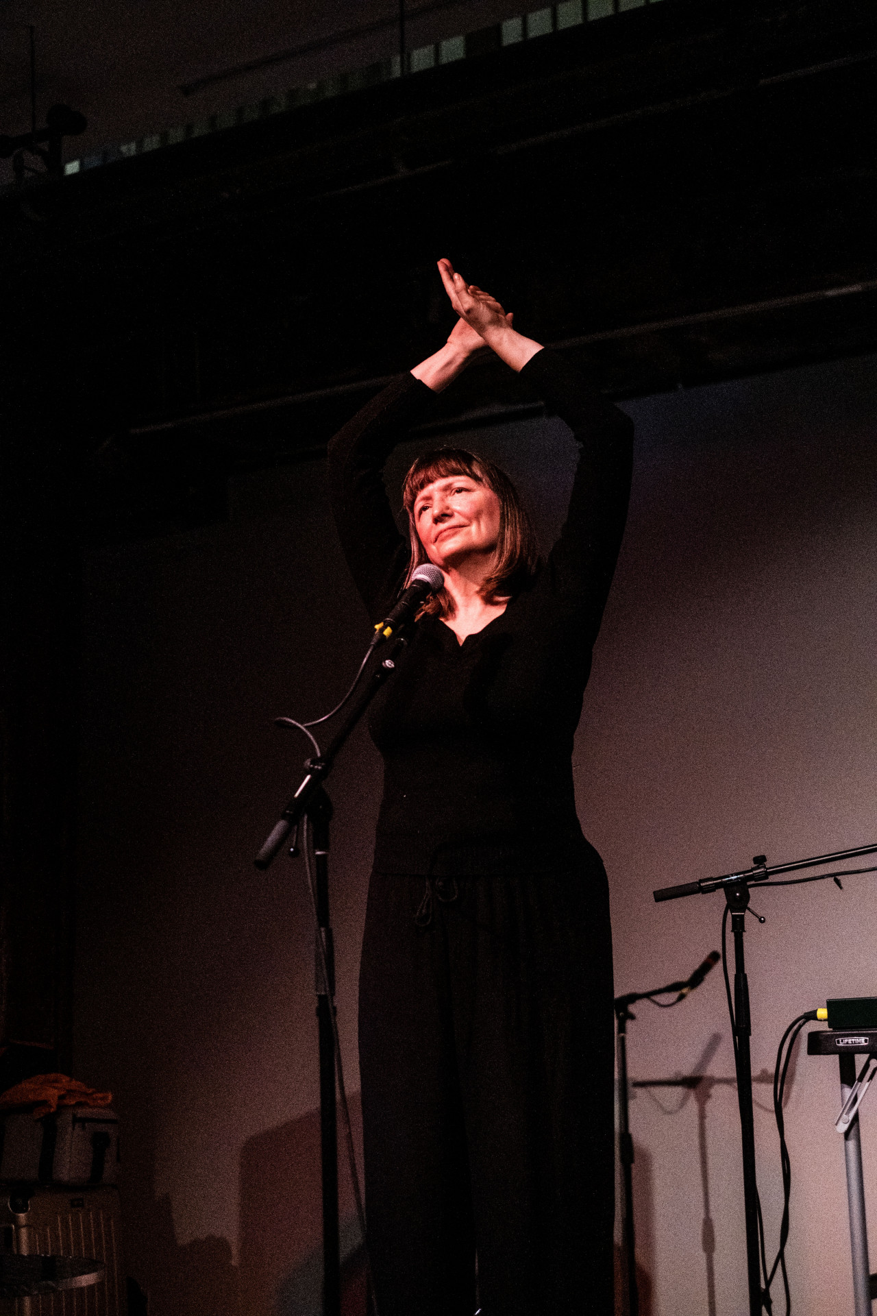 A figure dressed in black stands behind a microphone and stretches their arms above their head.