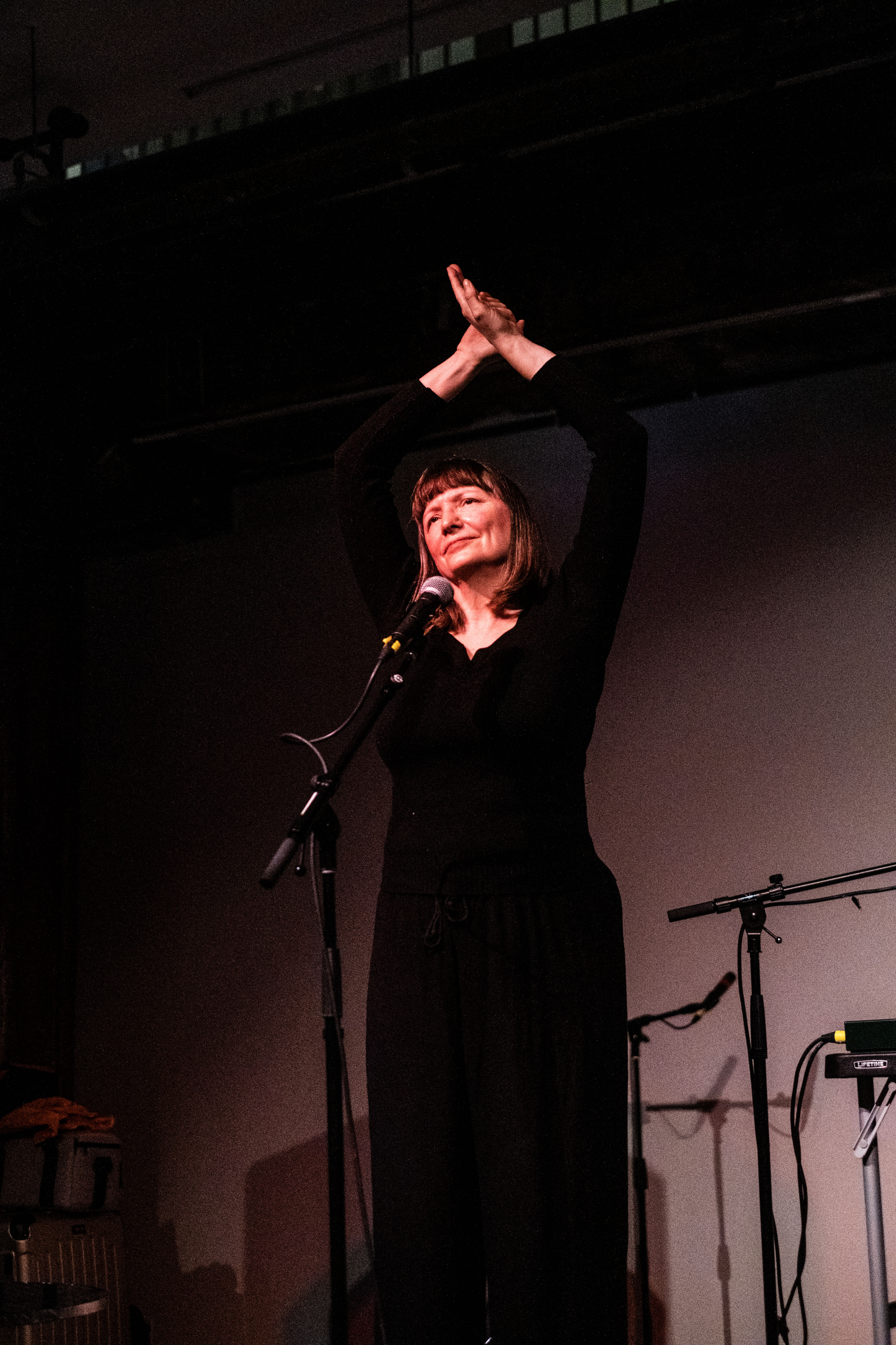 A figure dressed in black stands behind a microphone and stretches their arms above their head.