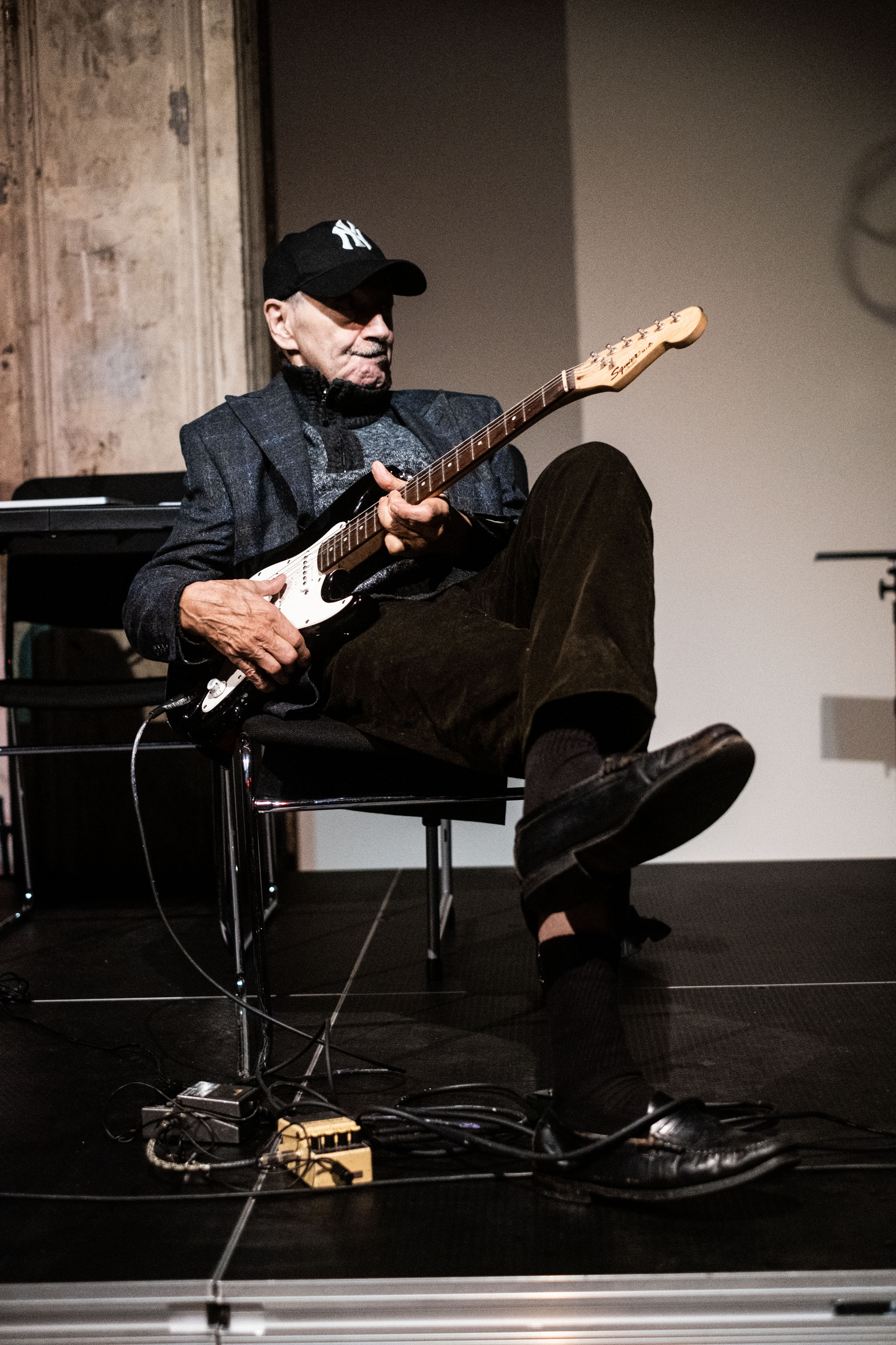 A figure wearing a baseball cap sits in a chair and plays the guitar, their legs crossed.