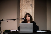 A figure sits behind a table with a laptop and a microphone in front of them.