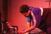 A figure wearing a blue shirt kneels down onto the stage, working on audio equipment. Red light is cast onto them.