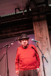 A figure in a red sweater and a black bowler hat stands behind three microphones, speaking into them.
