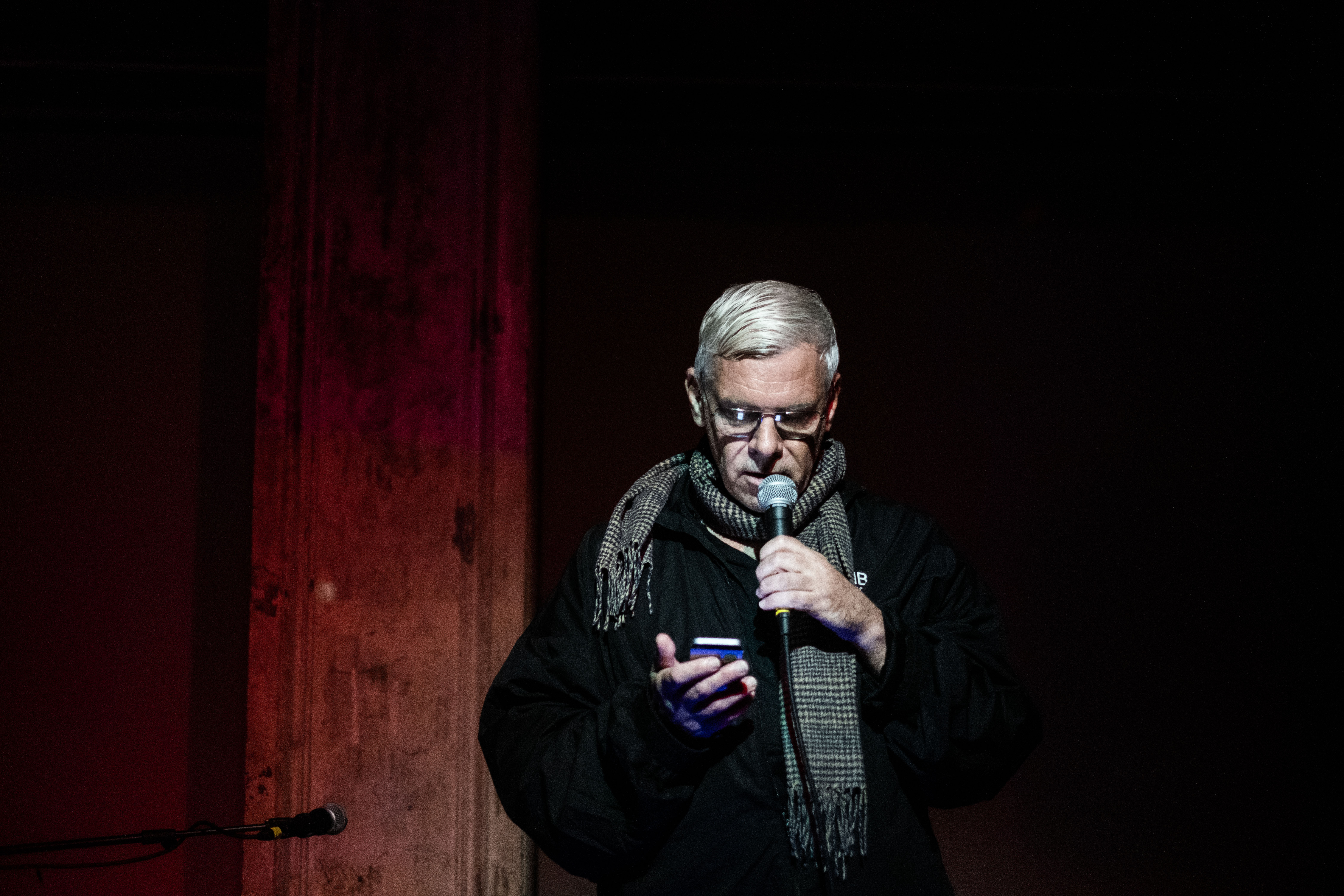 A figure reads into a microphone, glancing at their phone.