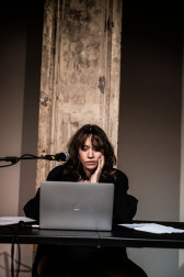 A figure sits behind a table, looking at a laptop screen, their face resting in their hand and a microphone placed next to their face.