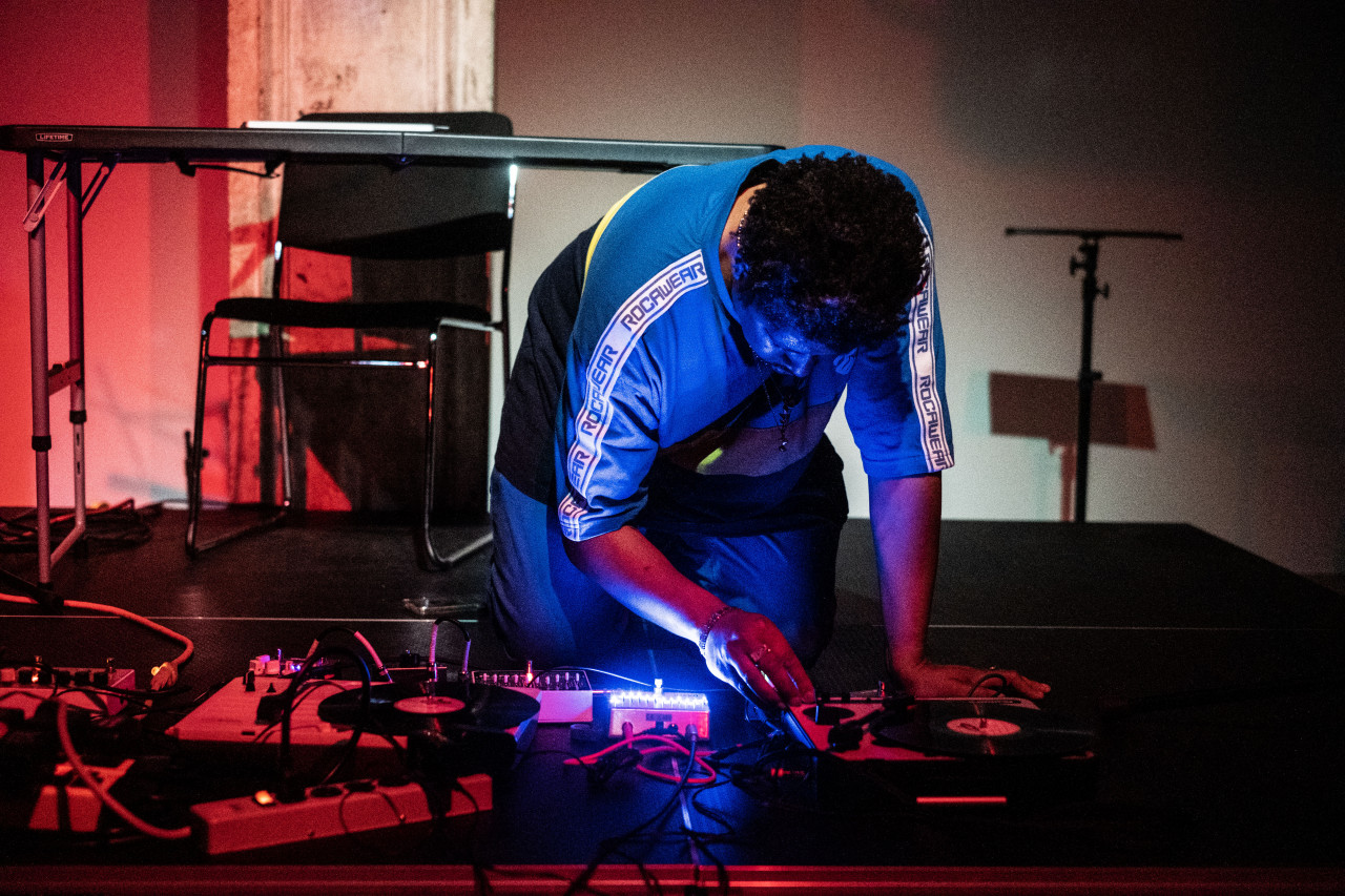 A figure kneels down on all fours on the stage, setting up audio equipment. Blue and red lights are cast onto them.