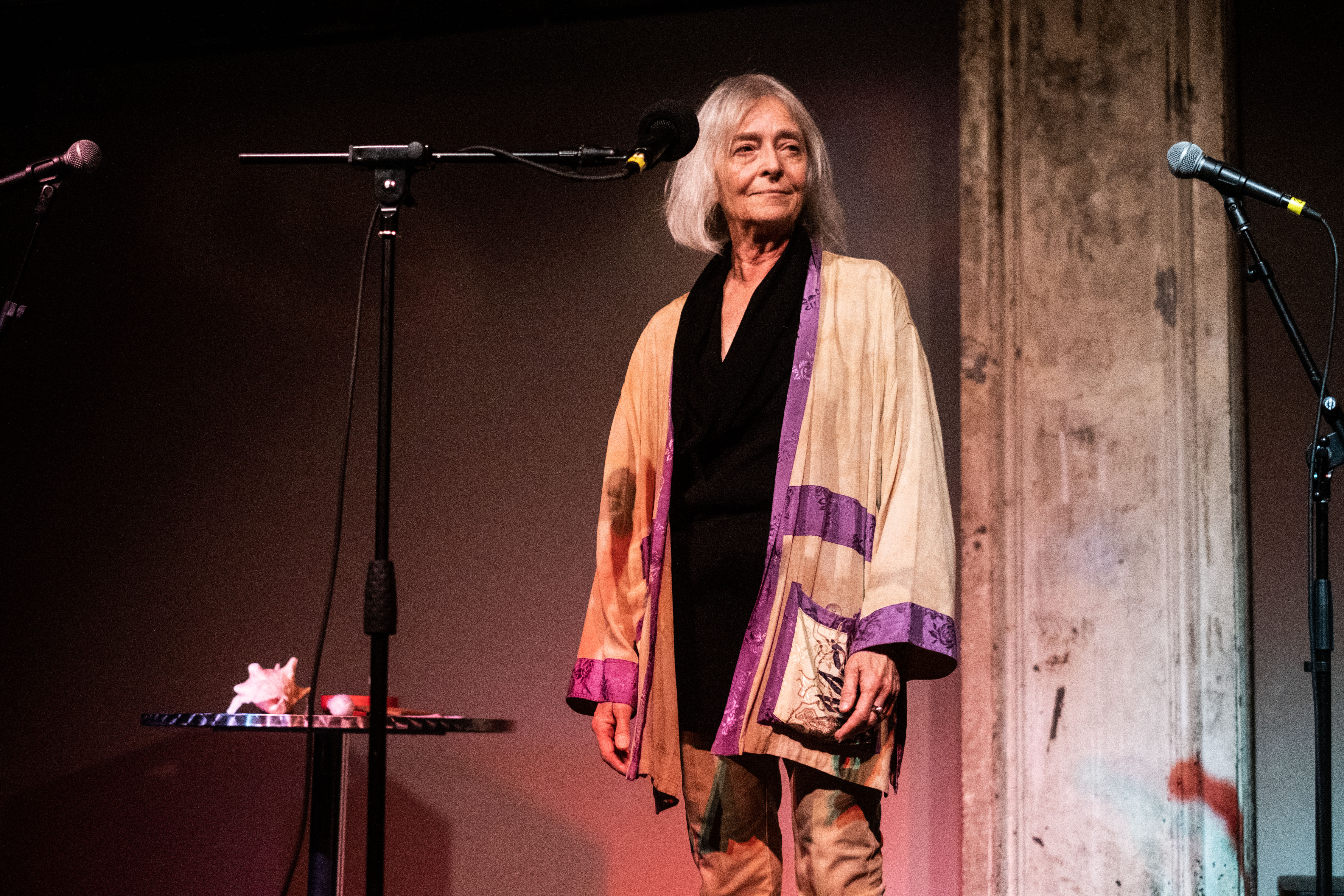 A figure stands behind a microphone, smiling and looking off into the audience.