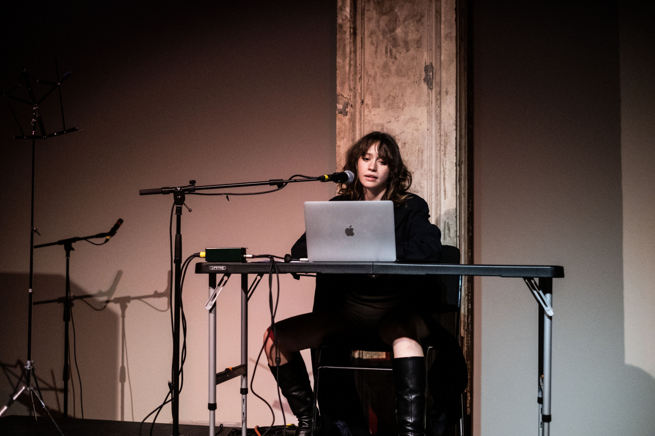 A figure sits behind a table, looking at a laptop, and speaking into a microphone.