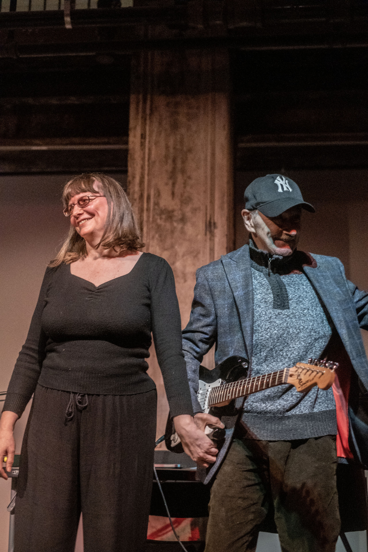 Two figures stand on stage next to each other, holding hands and smiling at the audience.