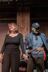 Two figures stand on stage next to each other, holding hands and smiling at the audience.