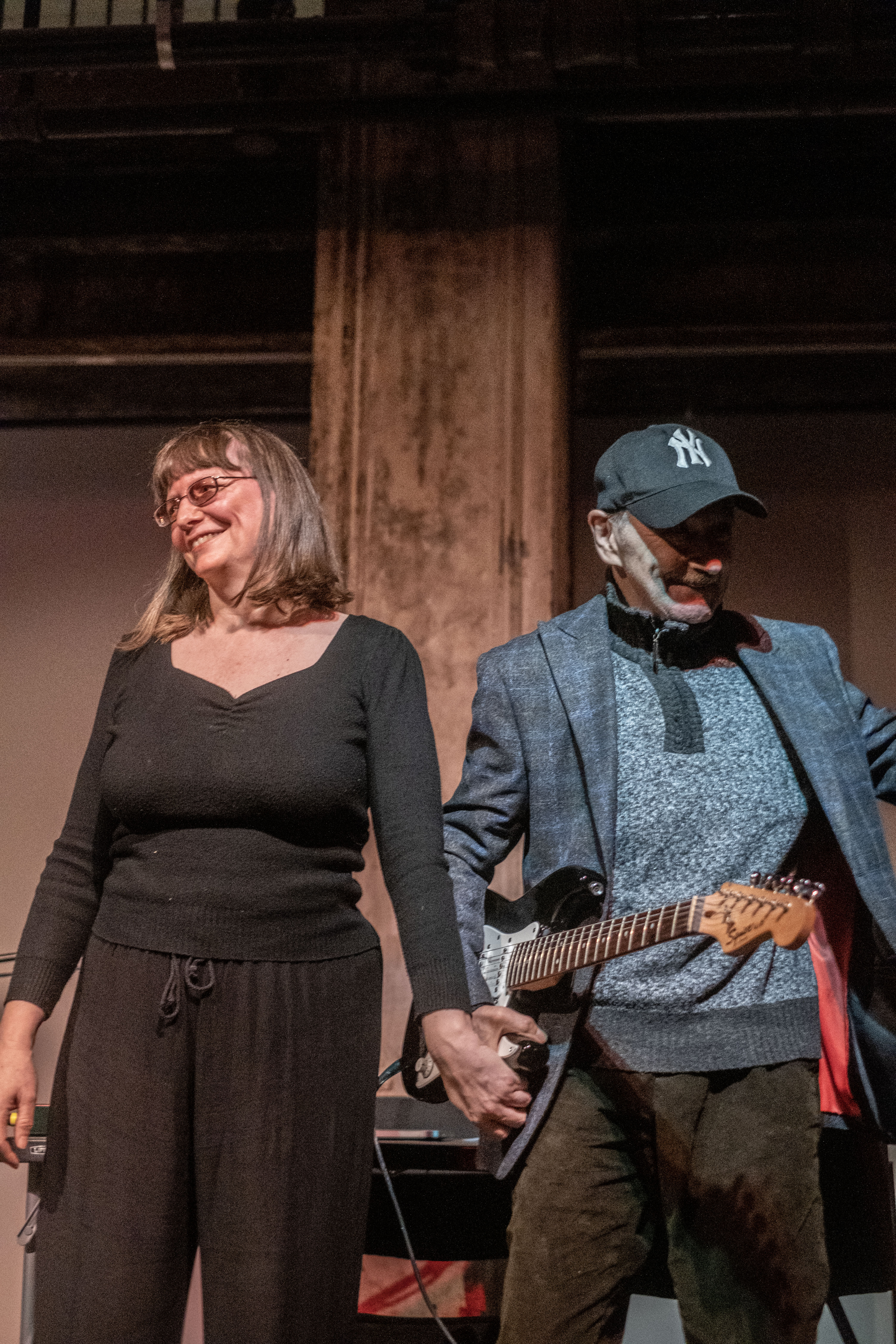 Two figures stand on stage next to each other, holding hands and smiling at the audience.