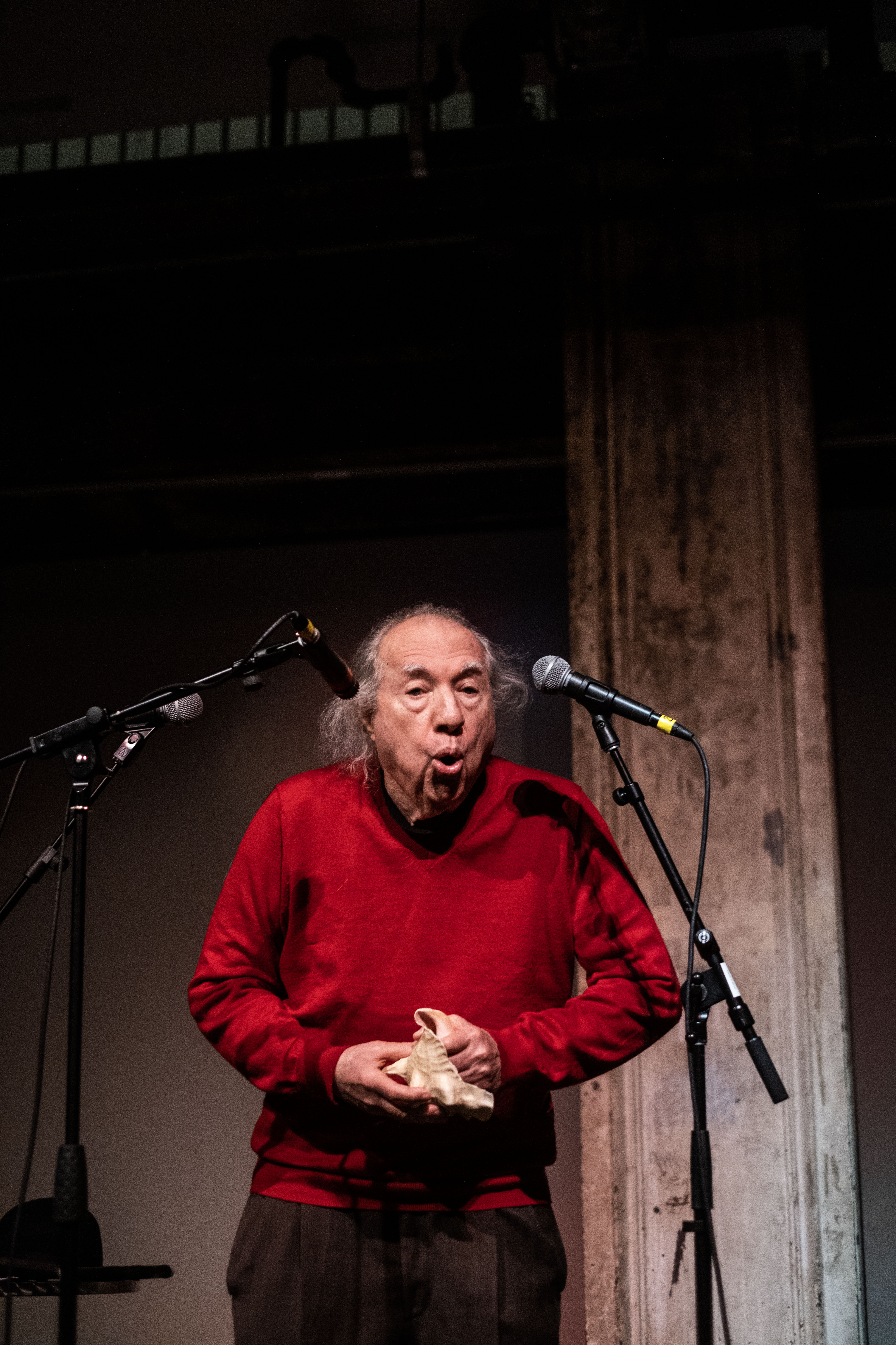 A figure in a red sweater stands on stage, with three microphones placed around them. They hold a white conch shell in their hands, their mouth is open.