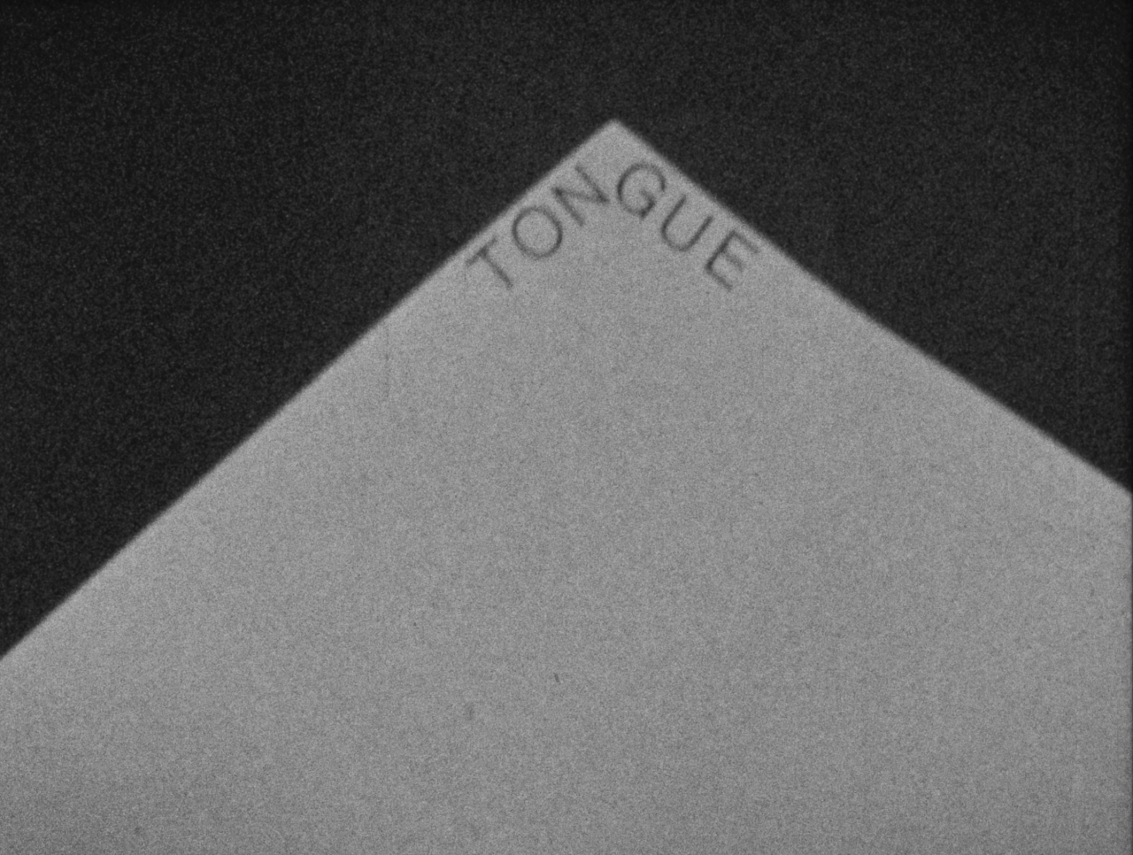 Black-and-white film still of a triangular shaped white surface against a black background. Along the edges of the triangle is text reading "TONGUE."