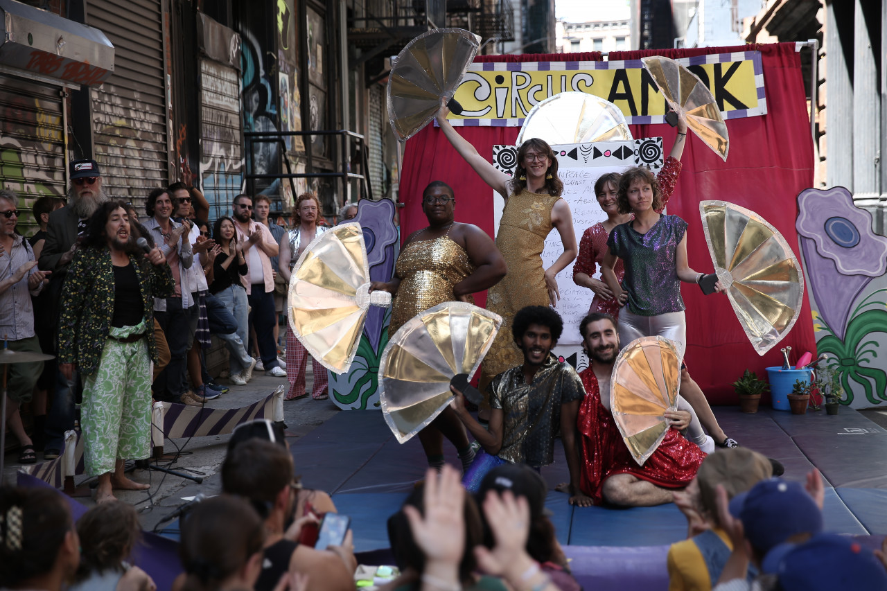 Various figures in glittery costumes pose in front of a circus backdrop, holding out open fans.