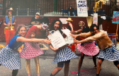 A color image depicts five figures performing on the street. The five figures are wearing blue or red American flag patterned skirts with different shirts. The center figure holds two shopping bags with the text 