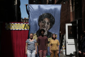 Three figures stand in front of a large banner bearing a portrait.