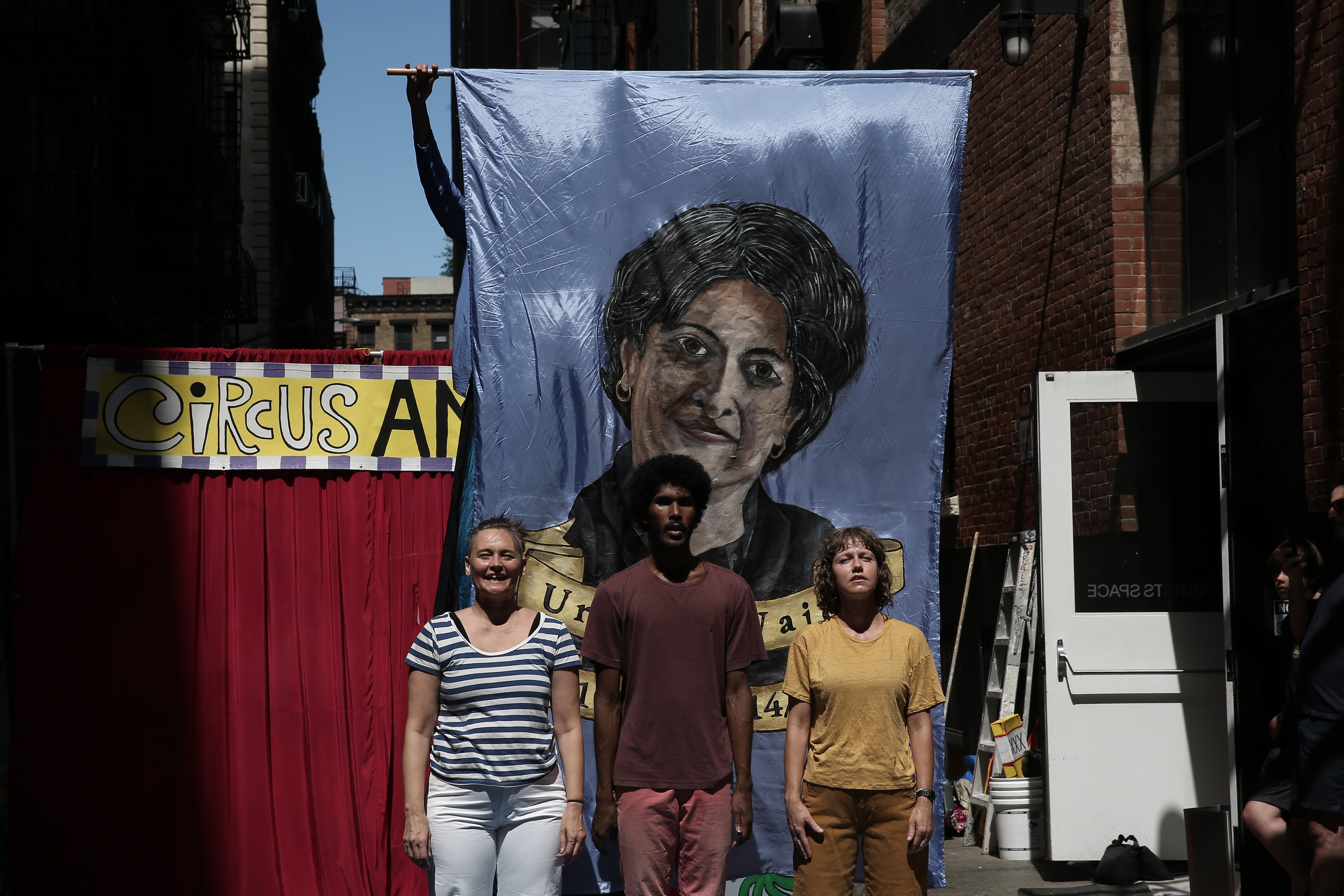 Three figures stand in front of a large banner bearing a portrait.