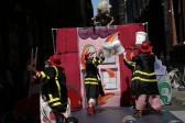 Three figures adorning firefighter costumes pass white buckets back and forth while being menaced from above by a figure wearing a wig.