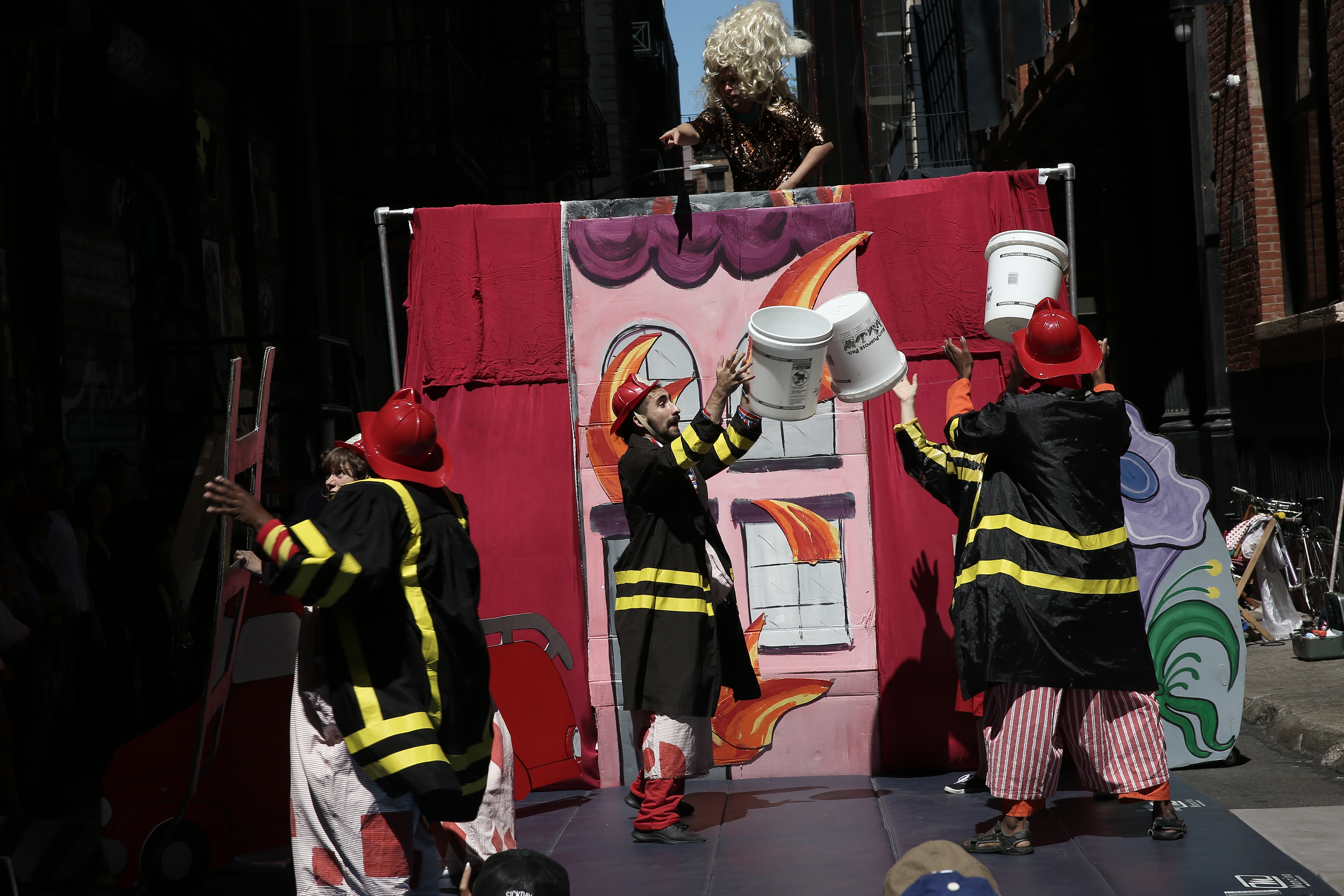 Three figures adorning firefighter costumes pass white buckets back and forth while being menaced from above by a figure wearing a wig.