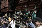 Five figures wearing ornate green outfits play instruments. In front of them, a crowd can be seen sitting down.