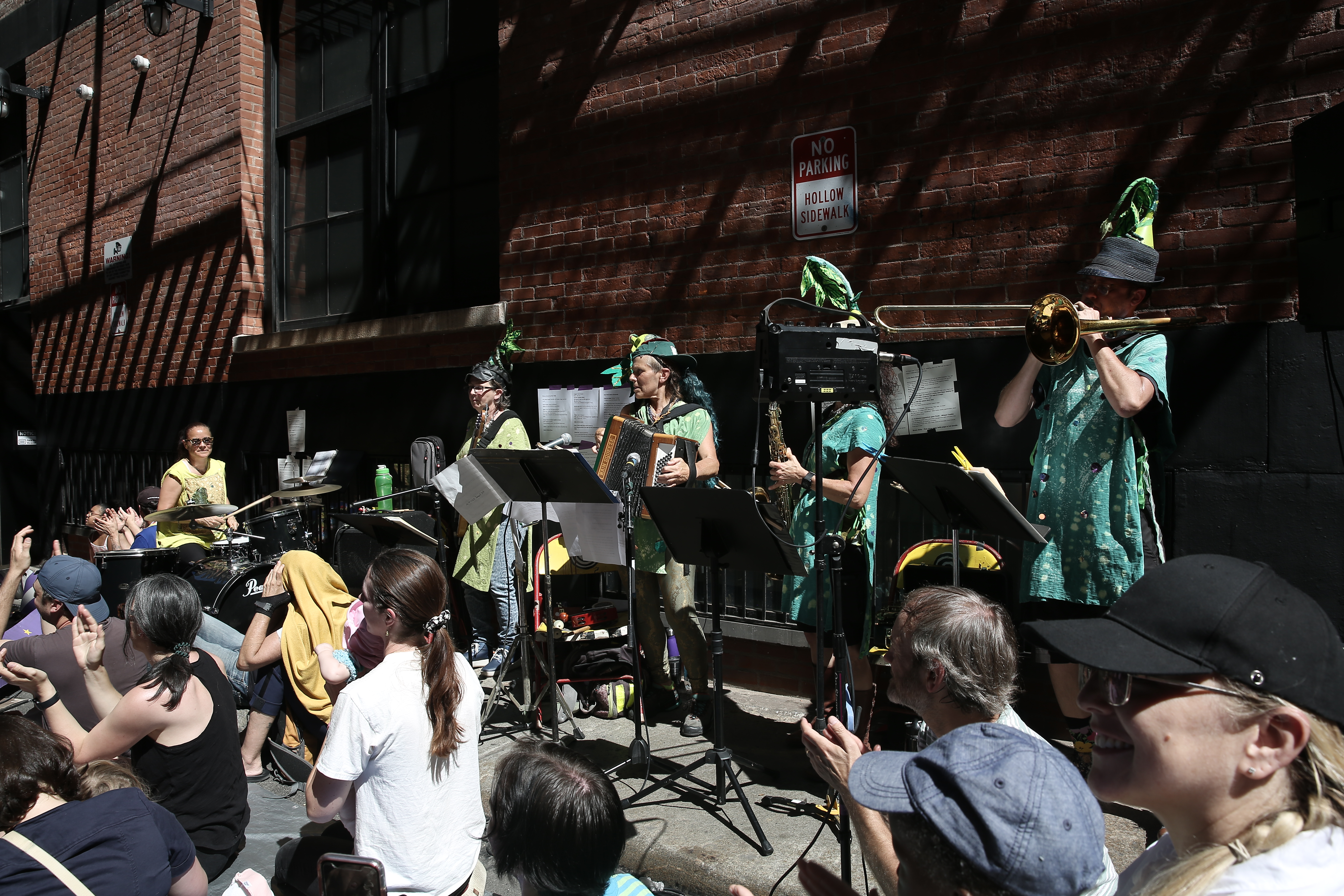 Five figures wearing ornate green outfits play instruments. In front of them, a crowd can be seen sitting down.