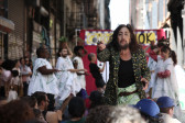 A figure points a microphone towards an audience, while various figures wearing white costumes and balancing plants on their heads can be seen just out of focus in the background.