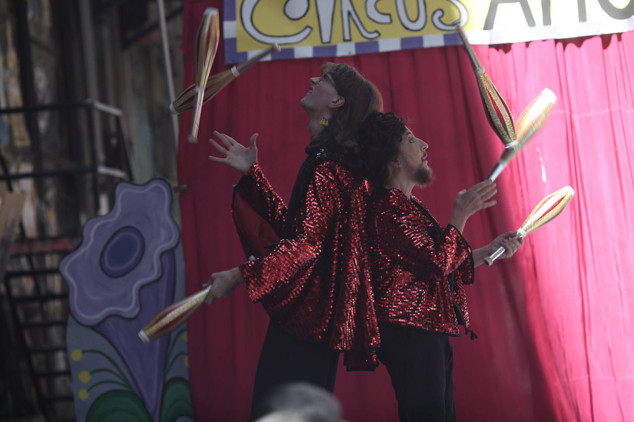 Two figures stand back to back, juggling clubs above their heads.