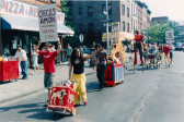 A color photograph of several figures marching in the middle of the street while holding up posters. One of the figures is wearing stilts, while others are pushing carts or instruments on wheels. From the sidewalk, various pedestrians look on.