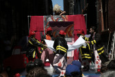 A group of figures dressed as firefighters spread out a tarp. Behind them is a cardboard set illustrated to look like a fire.