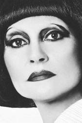 A black and white closeup of a person's face. They have blunt bangs and dark, dramatic eye makeup and lipstick.