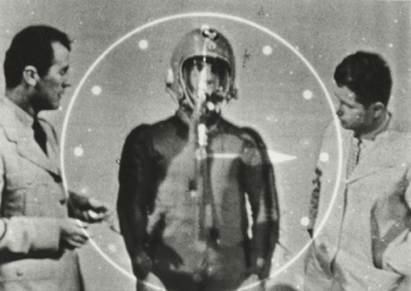 A black and white photo of three figures. The faint circular outline of a clock face surrounds the middle one, who is wearing a suit and a helmet.