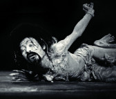 A black and white image of a man lying on the ground, his arms and legs splayed out as if in agony. His eyes are closed and his face is contorted.