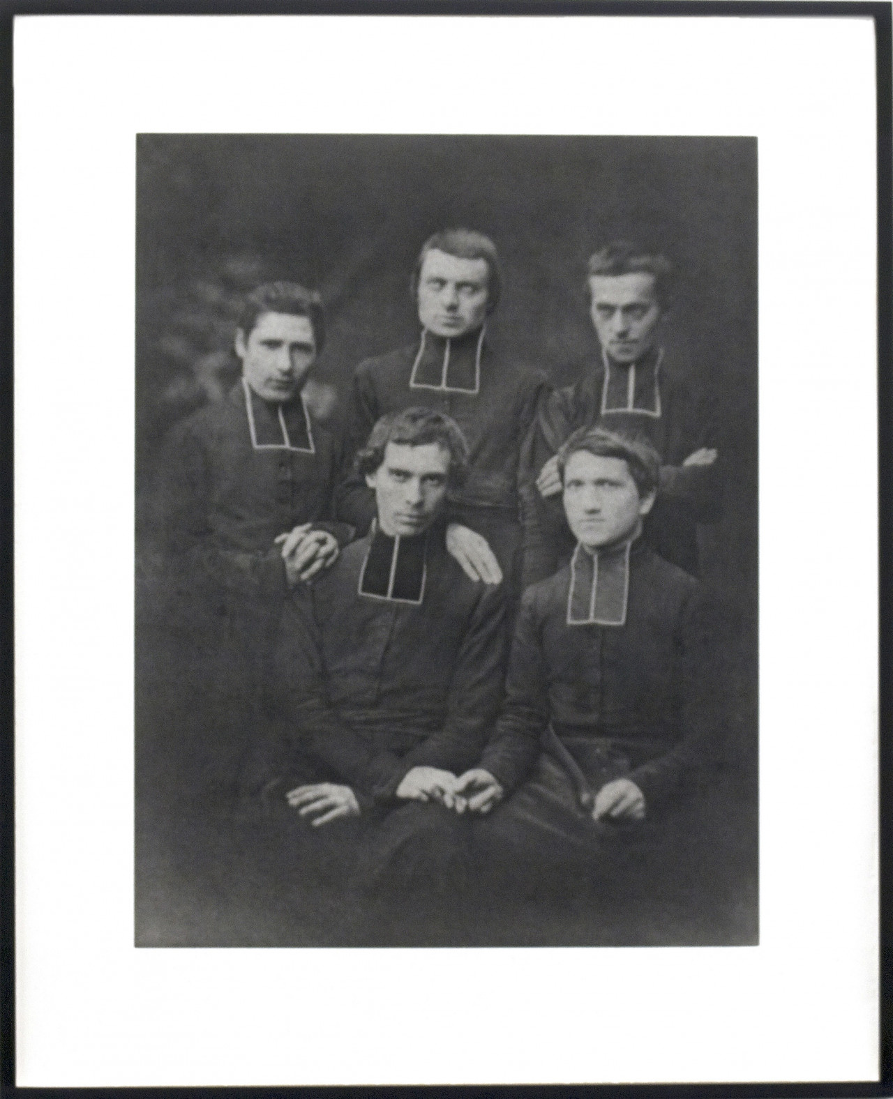 A vintage, framed black and white photograph of five priests dressed in all black stiffly pose before the camera for a portrait. The two men sitting closest to the viewer hold hands.