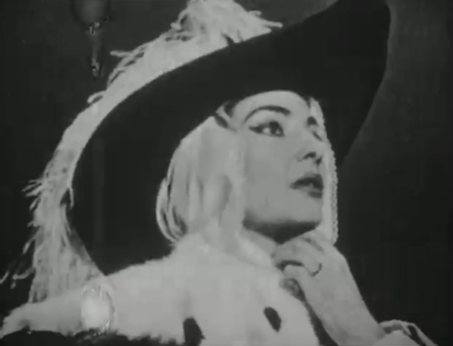 A black-and-white photo of a woman wearing a large black hat, holding her hand loosely to her neck, against a black background.