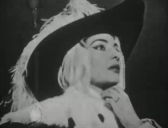 A black-and-white photo of a woman wearing a large black hat, holding her hand loosely to her neck, against a black background.