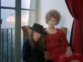 Two people sitting in front of a window looking towards the camera, one wearing a red dress and the other a black shirt and top hat.