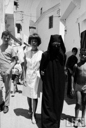 Black and white photograph of two women, one in a white dress and the other in a black niqab and sunglasses, walking arm in arm in a narrow street.