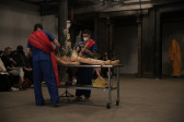Two figures wearing medical scrubs stand across from each other, with a table with a mannequin on it in the middle.