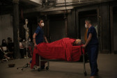 Two figures wearing medical scrubs and surgical masks move a table with wheels, on top of which a mannequin lies.