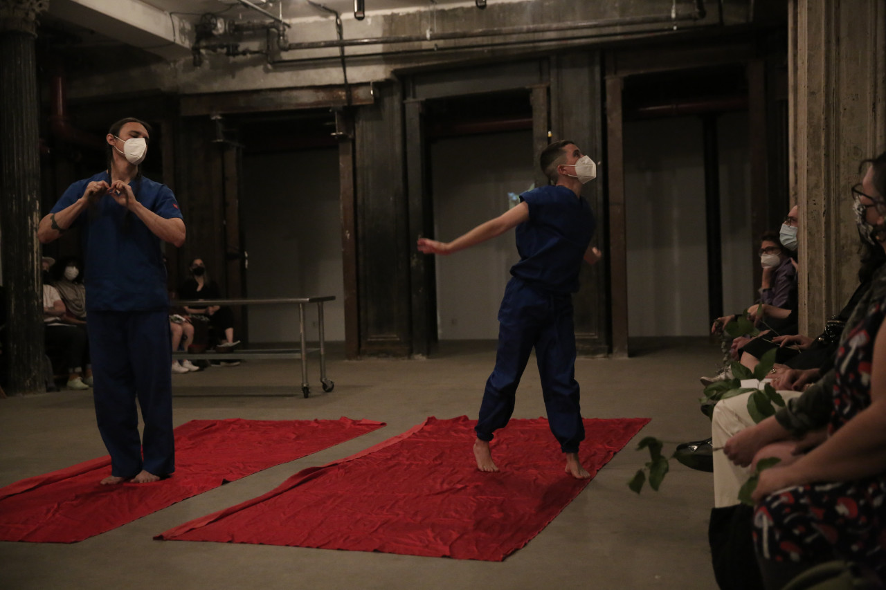 Two figures wearing medical scrubs stand on pieces of red fabric, stretching outwards toward the audience.