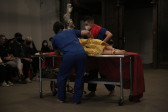 Two figures wearing medical scrubs move a table with wheels, on which a mannequin lies.